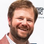 Smiling bearded man in a blazer and collared shirt against a branded event backdrop